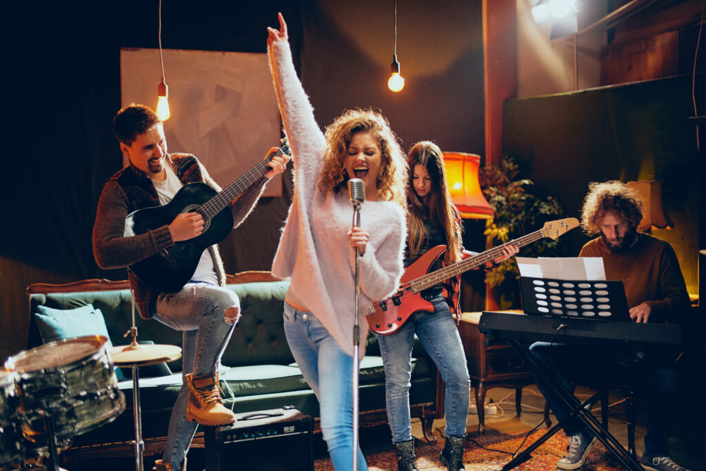 Woman with curly hair holding microphone and singing while man in background playing acoustic guitar